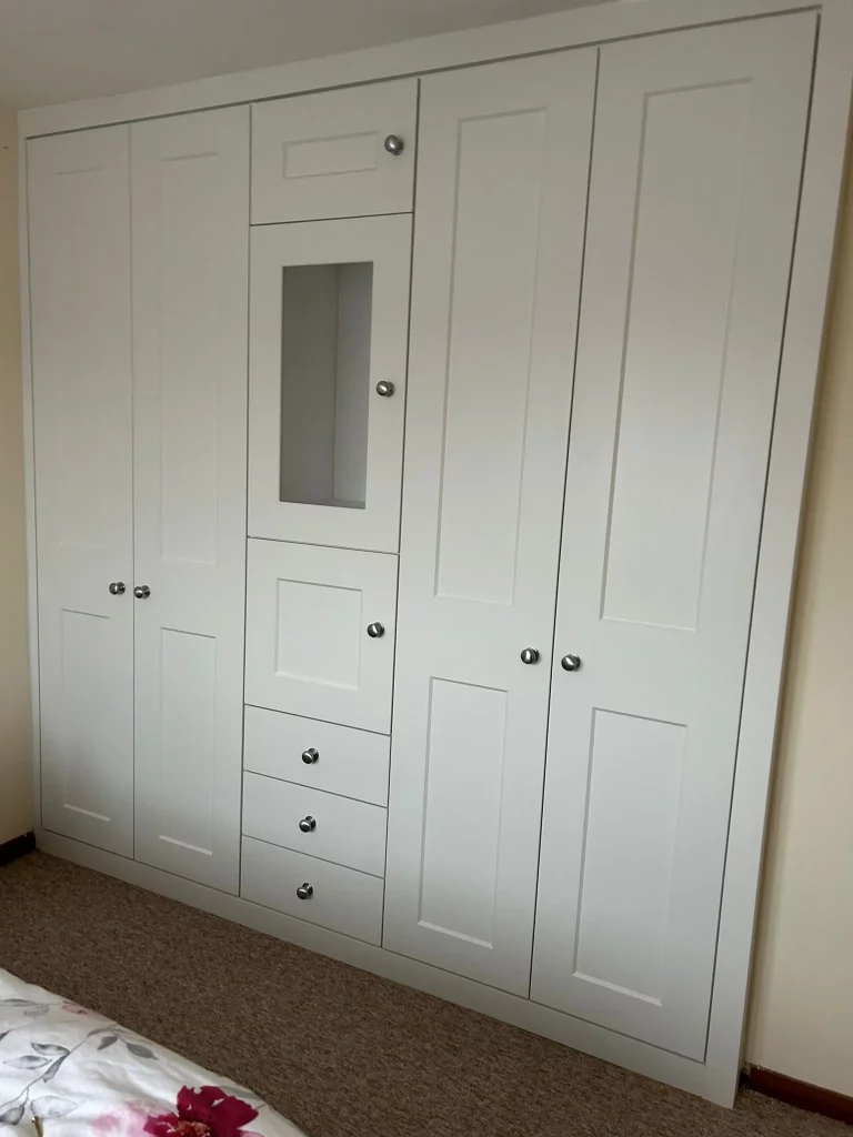 White built-in wardrobe in a bedroom, featuring multiple doors and drawers with silver knobs. The design is sleek and modern, adding a tidy feel.